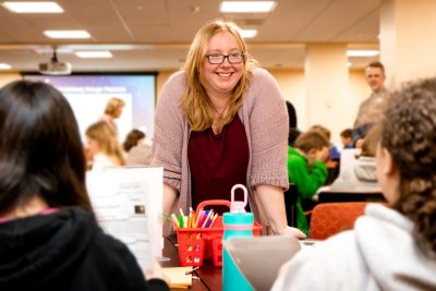 A woman leans over a table and a caddy of colorful markers to talk to students.