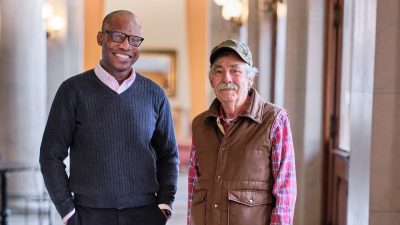 Two men of different races, ages, and dress stand together in a hallway.