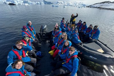 People wearing matching winter coats and life preservers sit in two inflatable zodiac boats, with icebergs behind them. 