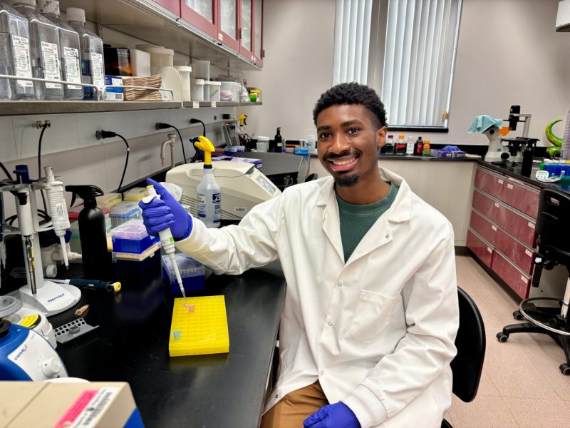 A smiling young man sitting in a lab holds a dropper.