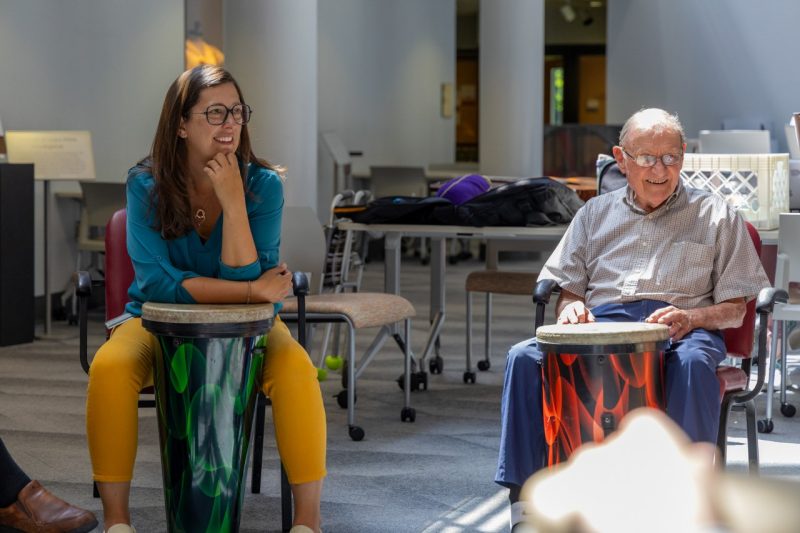 A woman with long dark hair and glasses is wearing a bright blue top and yellow pants. She is seated, leaning against a conga drum. Beside her is a man with gray hair and glasses, wearing a short sleeve light-colored button and dark blue jeans. He is smiling and playing a conga drum.