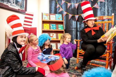 A woman dressed all in black with a giant red bow tie and a red and white striped top hat reads to a group of children.