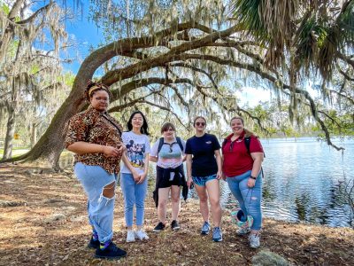 Five women pose for a photo in front of a live oak tree arched over the water of a lake.