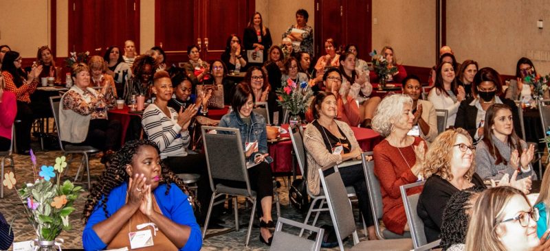 A roomful of women sitting at conference tables applaud.