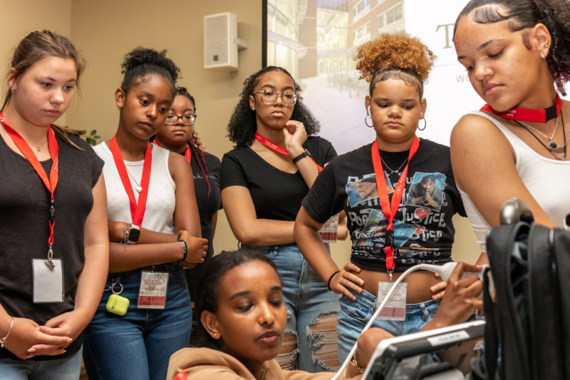 A young woman crouches down behind a teen girl sitting on a table while other teen girls gather around behind her to watch.