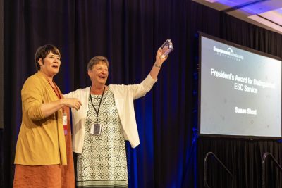 Two women stand together with one holding up a glass award. The award's name "President's Award for Distinguished ESC Service" is projected on a screen behind them.