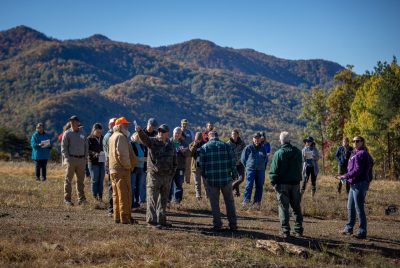 A group of people stand in the edge of a forested area with mountains in the background.