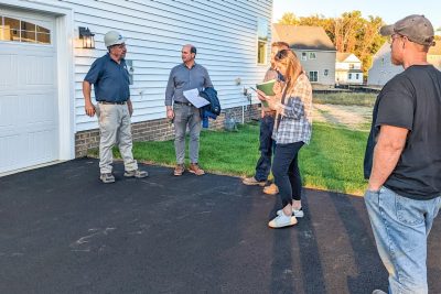 A group of people stand in the asphalt driveway of a new home listening to a man in a hardhat.