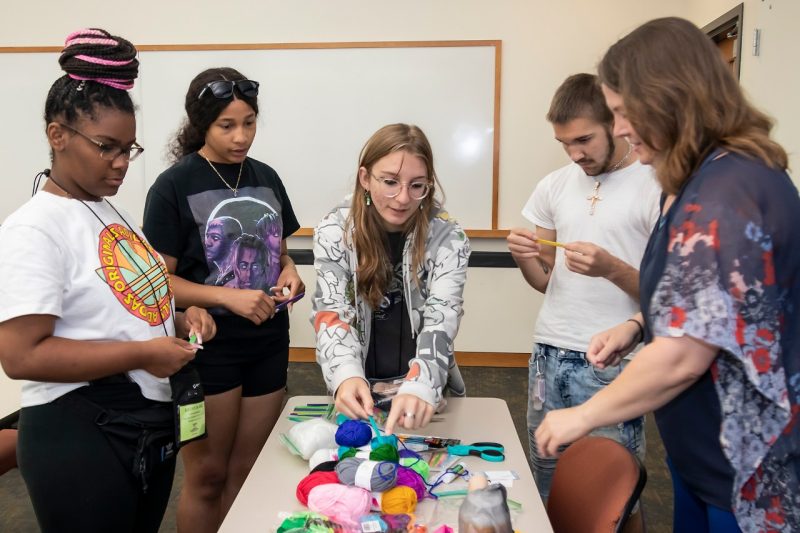 Four students and their teacher gather around a table covered in spools of colorful yarn and other crafting supplies.
