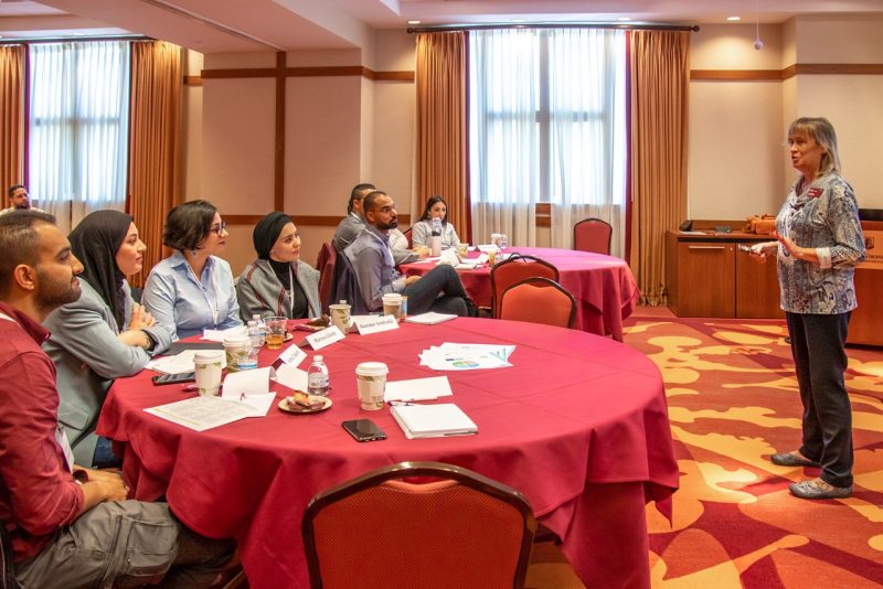 Men and women sit on one side of round tables while listening to a female professor speaking at the front of the room.