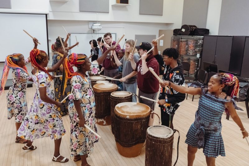 Women drummers from Rwanda teach their drumming techniques to a group of Virginia Tech students. The musicians and the students stand in the middle of a room, circling six big barrel drums, many of them holding drumsticks high in the air, ready to make contact with the drums. The drummers are are Black women with brightly colored long braids. They wear multi-colored patterned dresses. The students are dressed in sweatshirts and jeans and sweatpants.