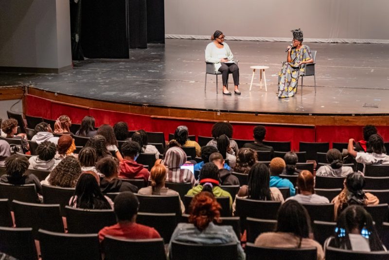 Artist and performer Kiki Katese talks with a student onstage in front of a large audience on students. Katese holds a microphone to her mouth. She is a Black woman wearing a multi-colored dress and multi-colored head dress and sits in a chair. The student with Katese onstage is also sitting in a chair. She is a black woman with glasses wearing a black headband, white sweater, and black pants.
