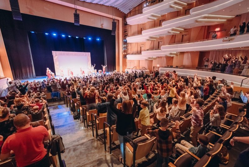 A theatre is filled with children standing up, many with their hands in the air as they watch a group of female Rwandan drummers perform onstage. The drummers have their hands in the air, also.