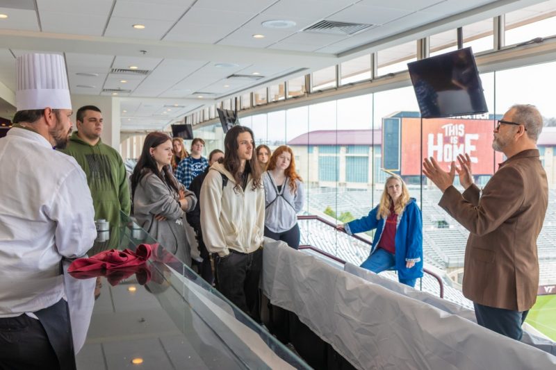 outreach-cac A group of high school students stand in a room overlooking the Virginia Tech football field listening to a man in a suit talk.