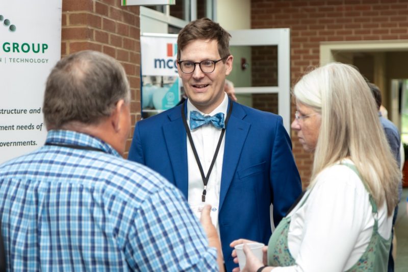 outreach-small-towns-conference A man in a blue suit jacket and blue bowtie talks with a man and a woman in a hallway.