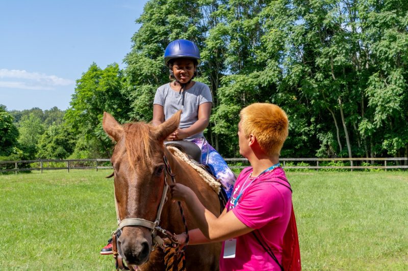 A young girl smiles sitting atop a brown horse, while a male counselor holds the bridle.