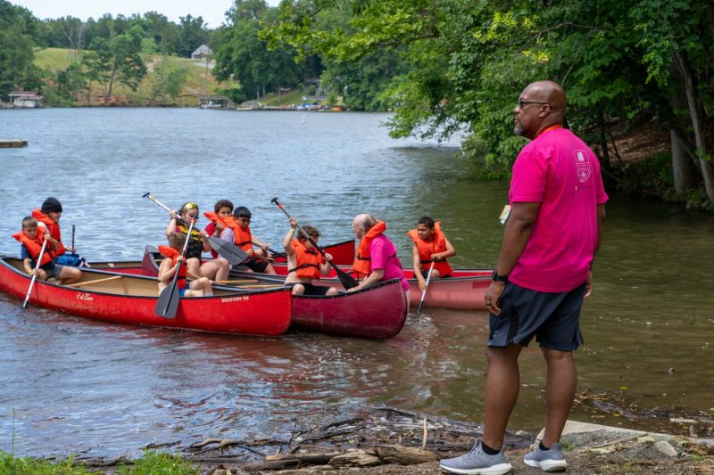 A man looks out at children paddling three red canoes in a lake.