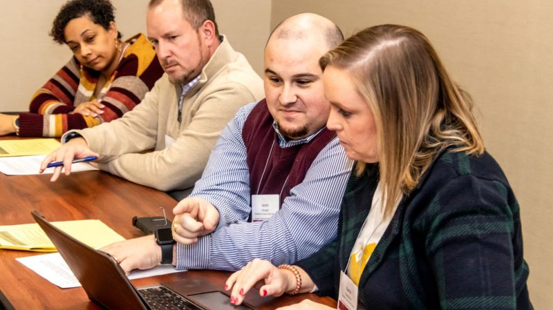 School Leaders Institute Four people sit at a wooden table. One man points at a laptop screen.
