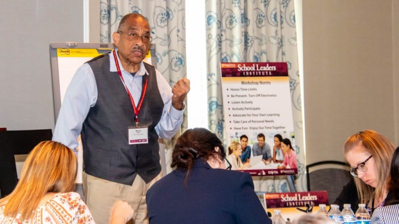 School Leaders Institute A man in a gray vest and wearing a lanyard speaks at the front of a class with a "School Leaders Institute" sign in the background.