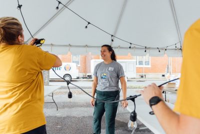 01_20240914_UtProsim_ElliTravis_CF_6481.jpg Elli stands under a large white tent with other volunteers as they work to hang string lights under the tent.