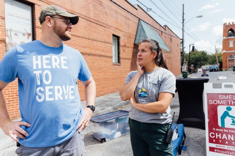 38_20240914_UtProsim_ElliTravis_CF_7647.jpg A man in a blue t-shirt that says "Here to Serve" speaks with a woman in a gray t-shirt outside a brick building.