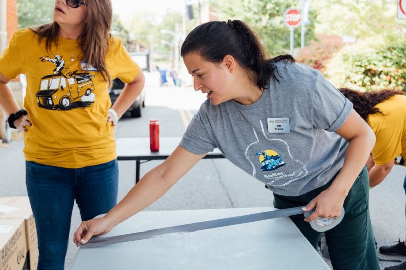 54_20240914_UtProsim_ElliTravis_CF_8115.jpg Elli marks out a folding table with duct tape. Two volunteers in yellow food truck rodeo shirts work in the background.