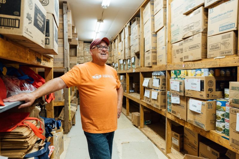 John Tarter, in an orange t-shirt, stands among shelves stacked with cardboard boxes and cans of donated food, to be given away at the Christmas Store.