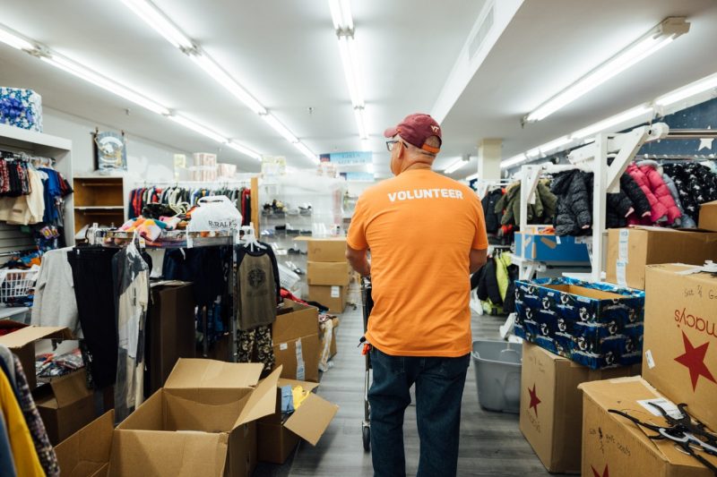 John Tarter walks amid boxes and racks of clothing at the store, wearing a t-shirt that reads "Volunteer" on the back.