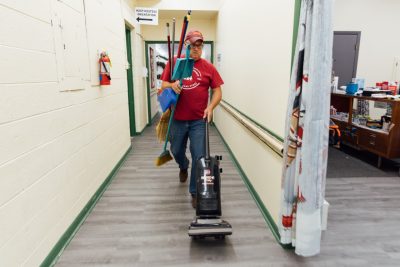 John Tarter walks down a hallway with a vacuum and several brooms.