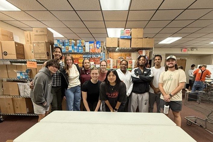 Eleven student leaders and Residential Well-being staff smile together in front of boxes of food in a warehouse, with other workers in the background.