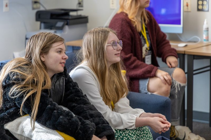 A group of three middle school girls are seated in a classroom, looking ahead and smiling.