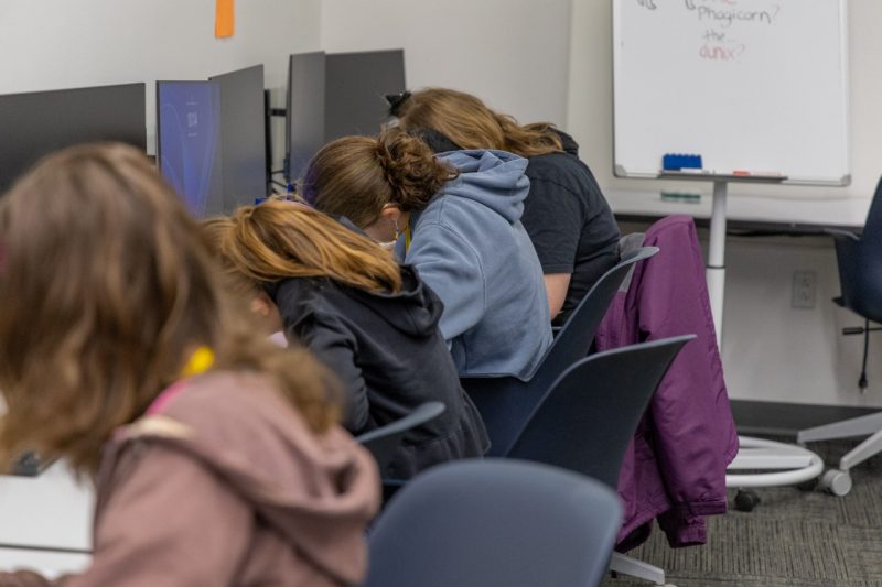 Four middle school students are seated in front of computers in a classroom. They are looking down, writing.