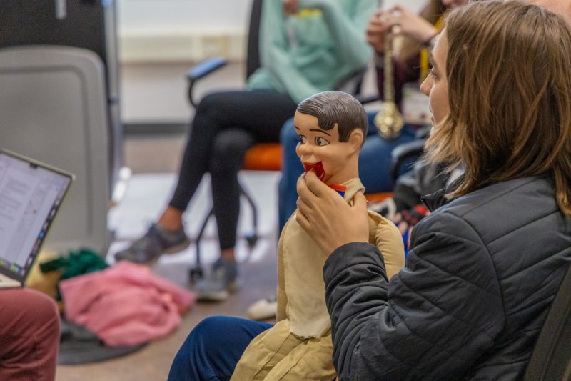 A student with long brown hair and a dark jacket is seated, holding a ventriloquy doll. Around him, other students are seated in a semicircle.