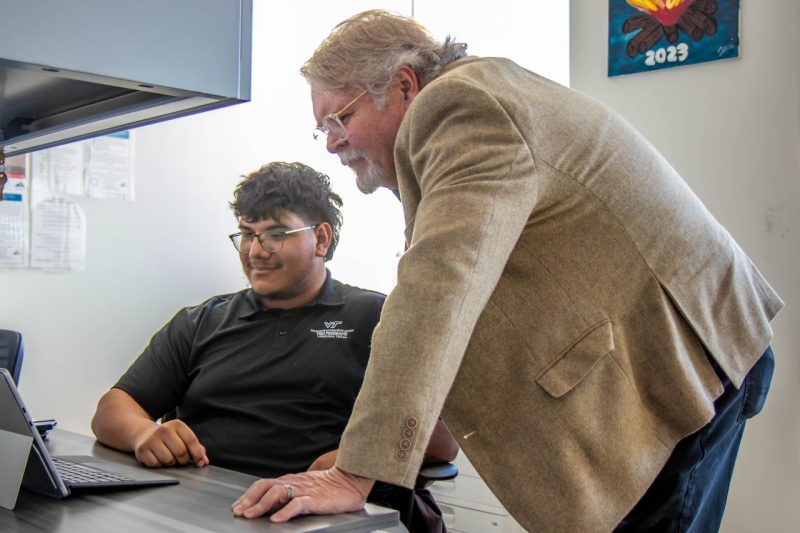A man in a suit jacket leans over a desk looking at a laptop screen with a seated student.
