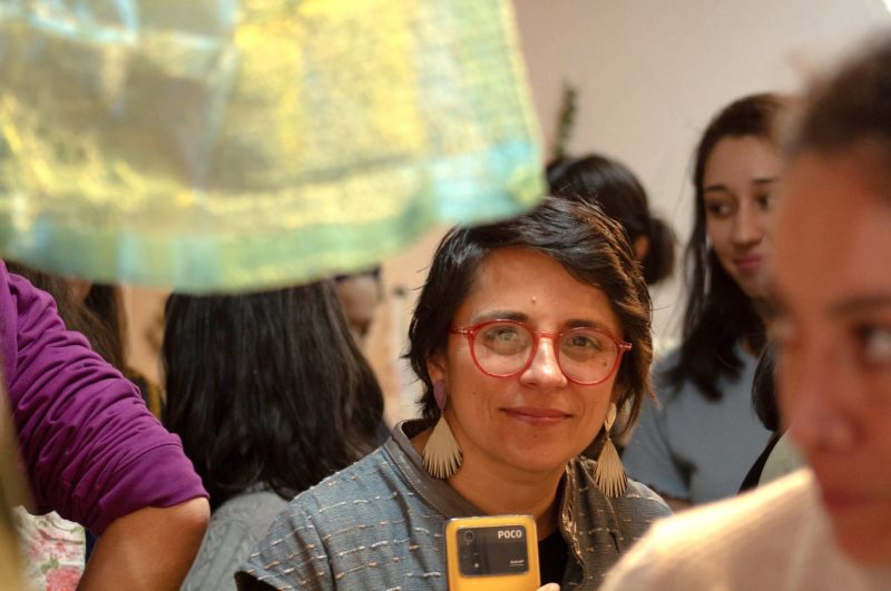 In a group of people Tania Perez-Bustos is centered in the frame, her attention is fixed on a piece of fabric that can be seen hanging in the foreground. 