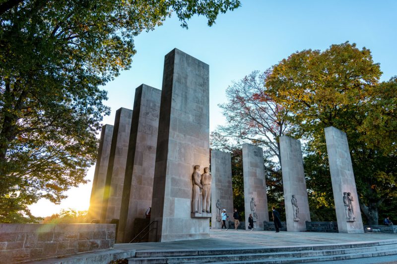 Stone pylons with the sun setting behind them and fall colors on the trees around them.