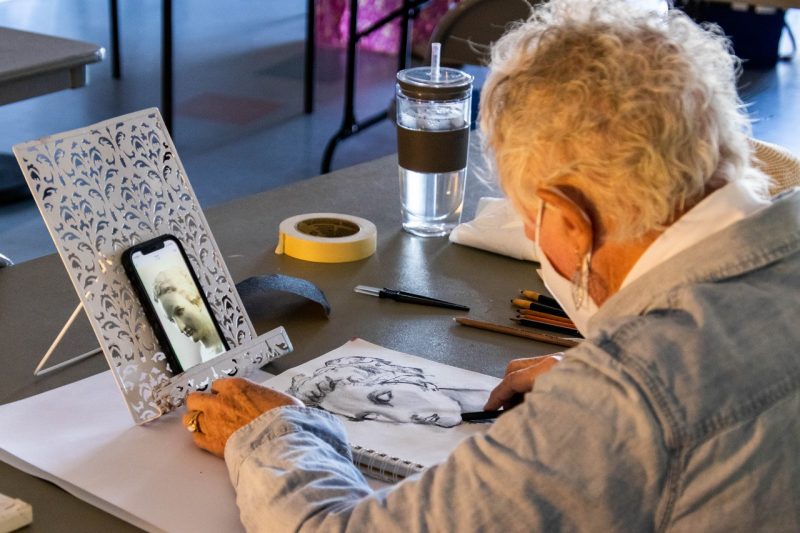 A woman draws with charcoal on an art pad while looking at a photo of a marble statue on her propped up cellphone.