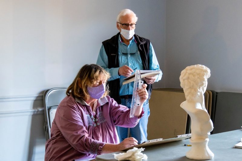 A woman sits holding a ruler up vertically toward a marble bust while a man studies the light on the statue from where he's standing to one side.