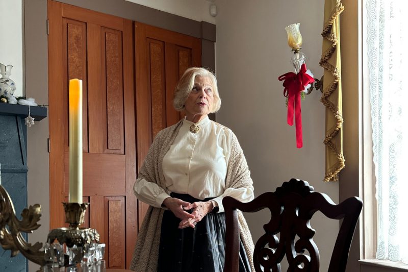 A docent dressed in a long skirt, white shirt, and tan shawl talks in the dining room.