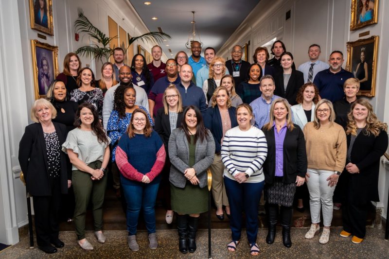 About 35 participants pose for a group photo on a set of stairs in a hallway.