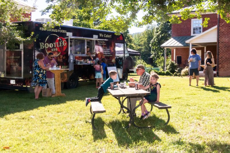 A family sits at a picnic table near a food truck with a old brick house in the background.