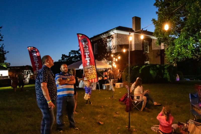 People talk and sit in chairs under a string of white lights