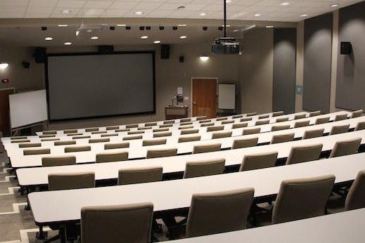 Tiered lecture hall with tan chairs and white tables that stretch across the room. 