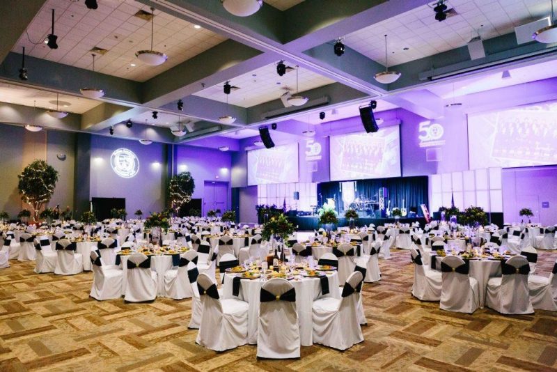 A grand ball room with purple lighting at the front and round tables with white linens.