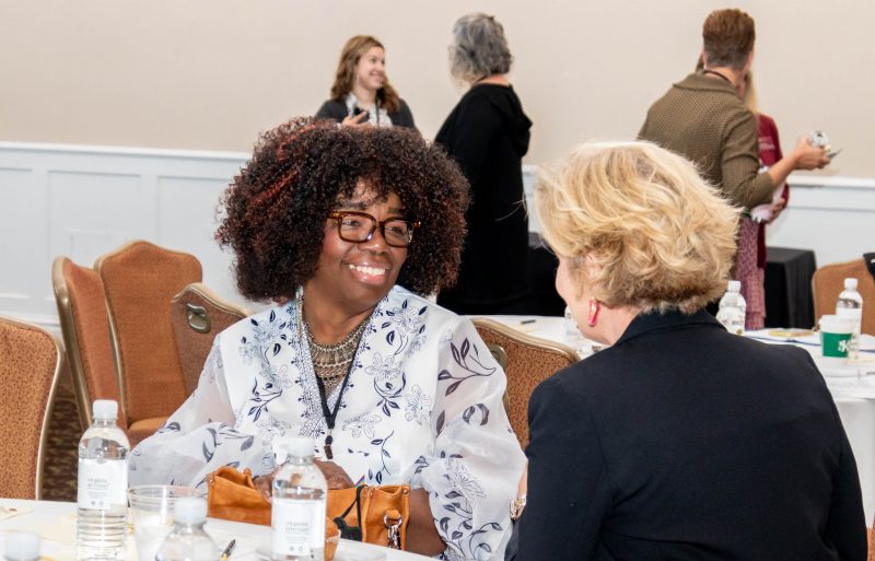 Kim B Miller, a Black woman with curly dark brown hair and glasses, wearing a black and white floral print dress and silver and black necklace, speaks with a summit attendee with short blonde hair, wearing a black blazer and red earrings. Several summit attendees can be seen in the background