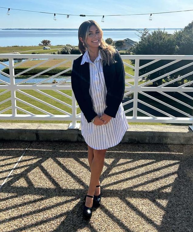 a young woman with brown and blonde hair, wearing a black and white pin striped dress and black cardigan poses in front of a white wooden railing with the James River visible in the background