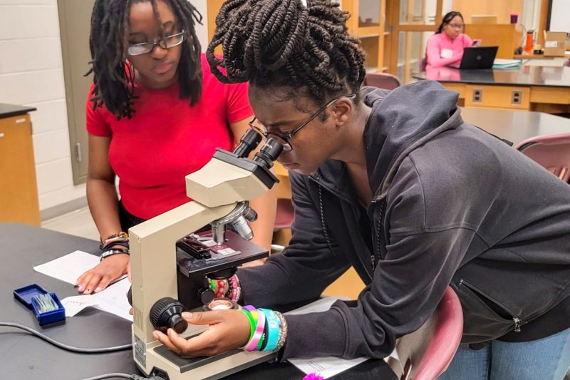 A teen girl leans over and looks into a microscope in a lab.