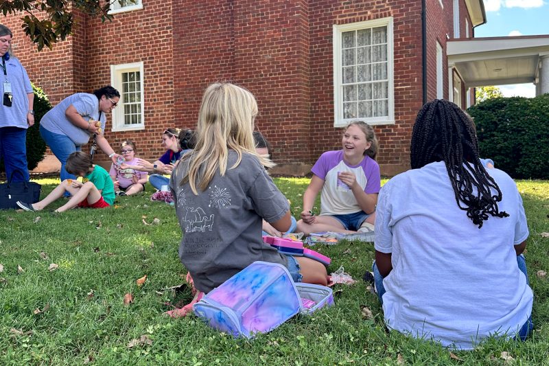 Three girls sit in the lawn eating their lunch next to a brick historic home.